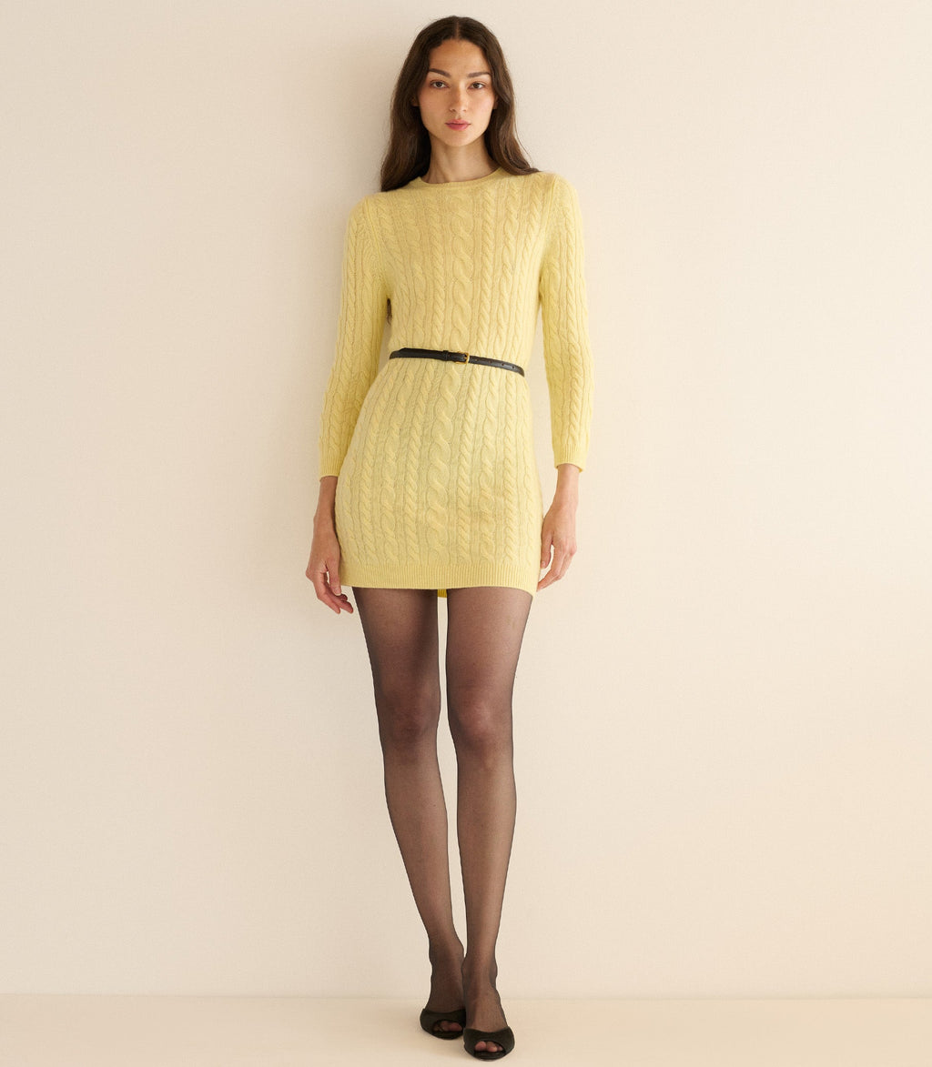 Woman wearing a yellow cable knit dress with a black belt against a beige background