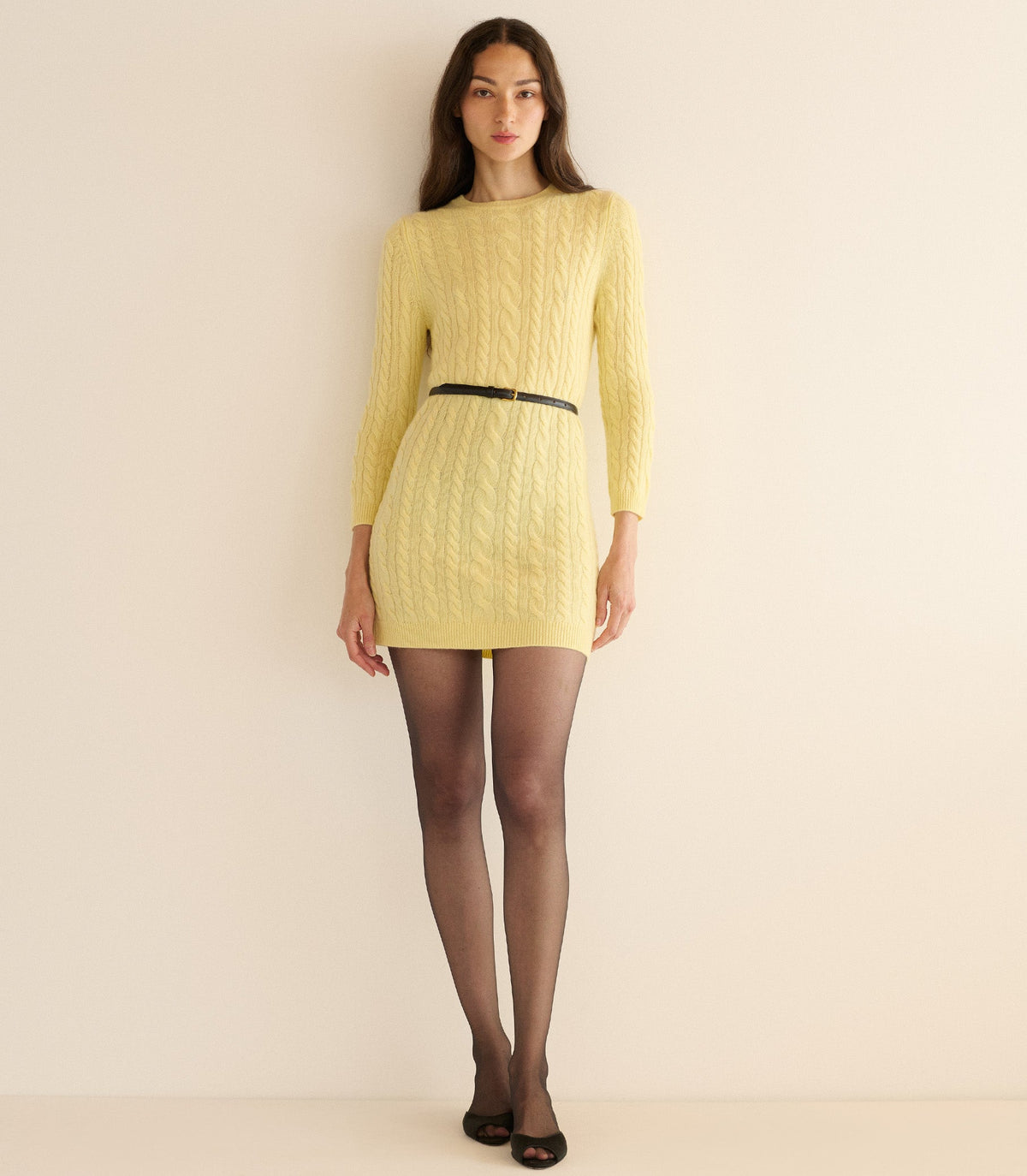 Woman wearing a yellow cable knit dress with a black belt against a beige background