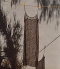 A sheer, dark brown lace slip dress hanging on a clothesline against a textured wall with tree shadows cast across it.