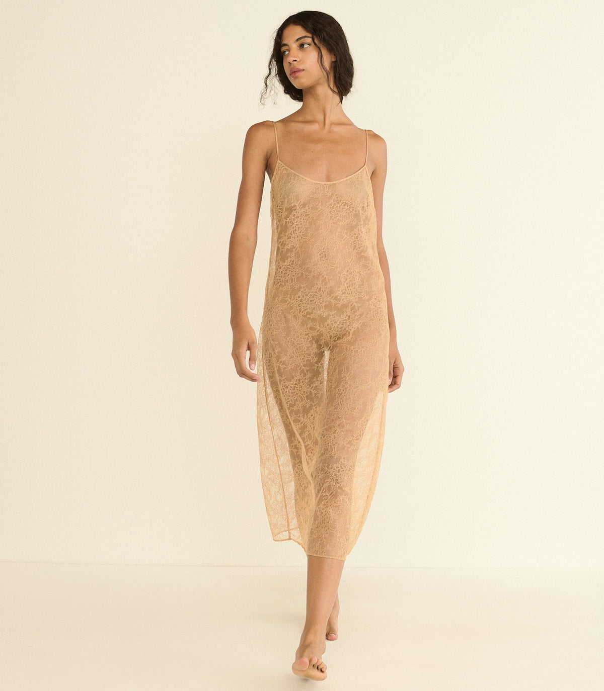 A woman modeling a sheer, textured slip dress in a light beige or tan color, standing against a solid, warm off-white studio background.