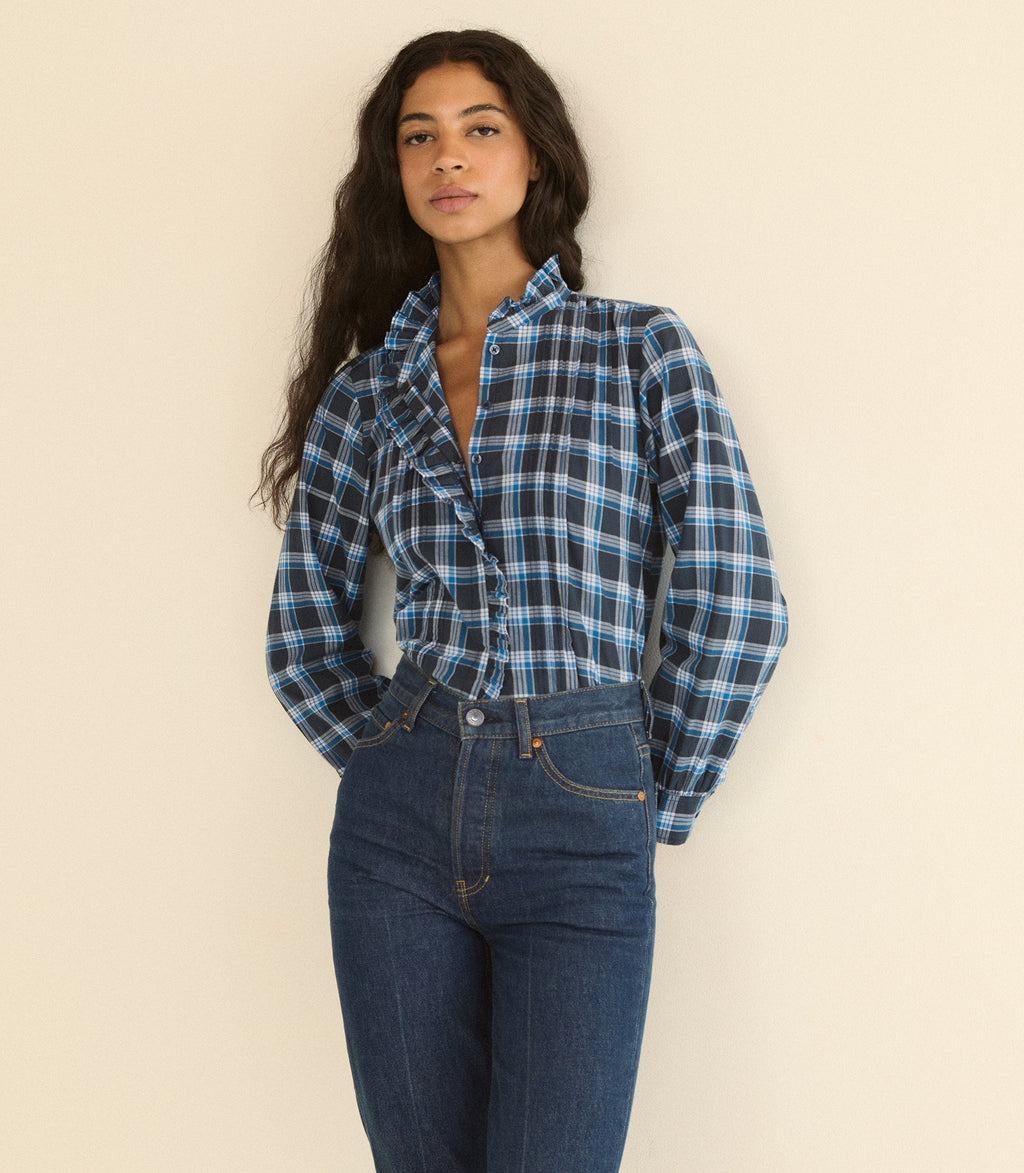 A woman wearing a blue and black plaid button-up shirt tucked into dark blue jeans, standing against a plain beige background.