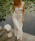 A woman wearing a long cream lace dress stands barefoot on a stone ledge beside a pond covered with lily pads.