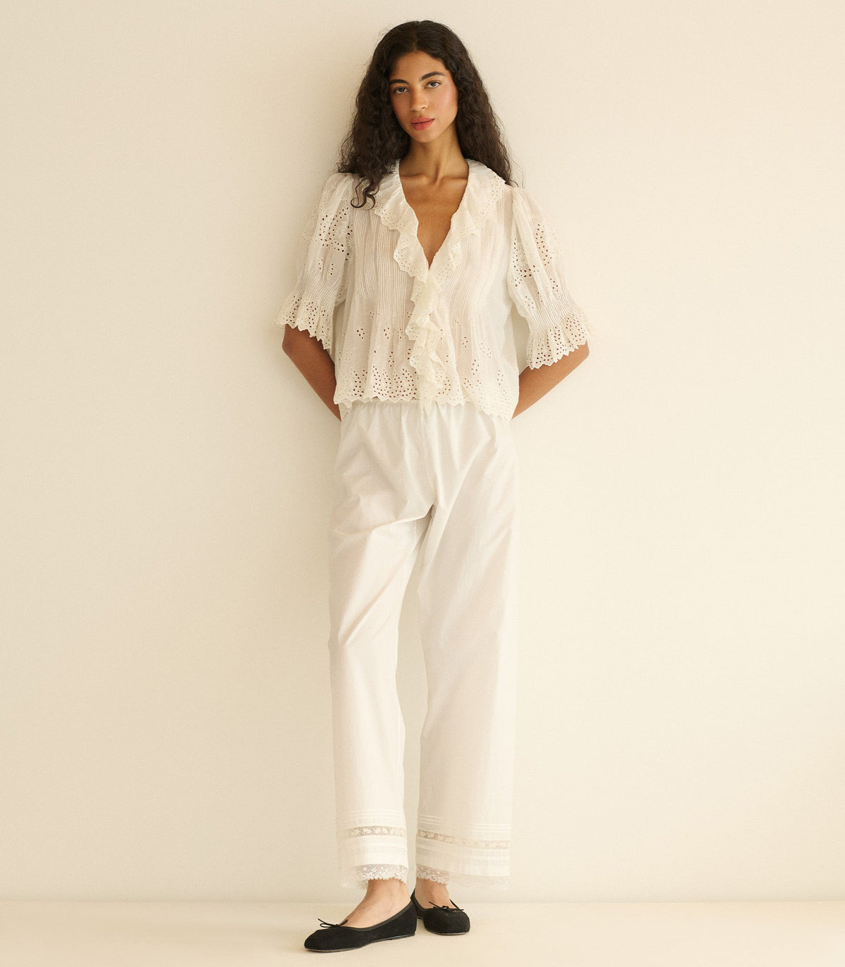Woman wearing a white eyelet blouse and white pants with lace details on a plain background
