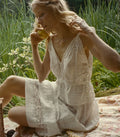 A woman sitting outdoors on a picnic blanket in a grassy area, wearing a loose white sleeveless top, white skirt, and holding a yellow fruit.
