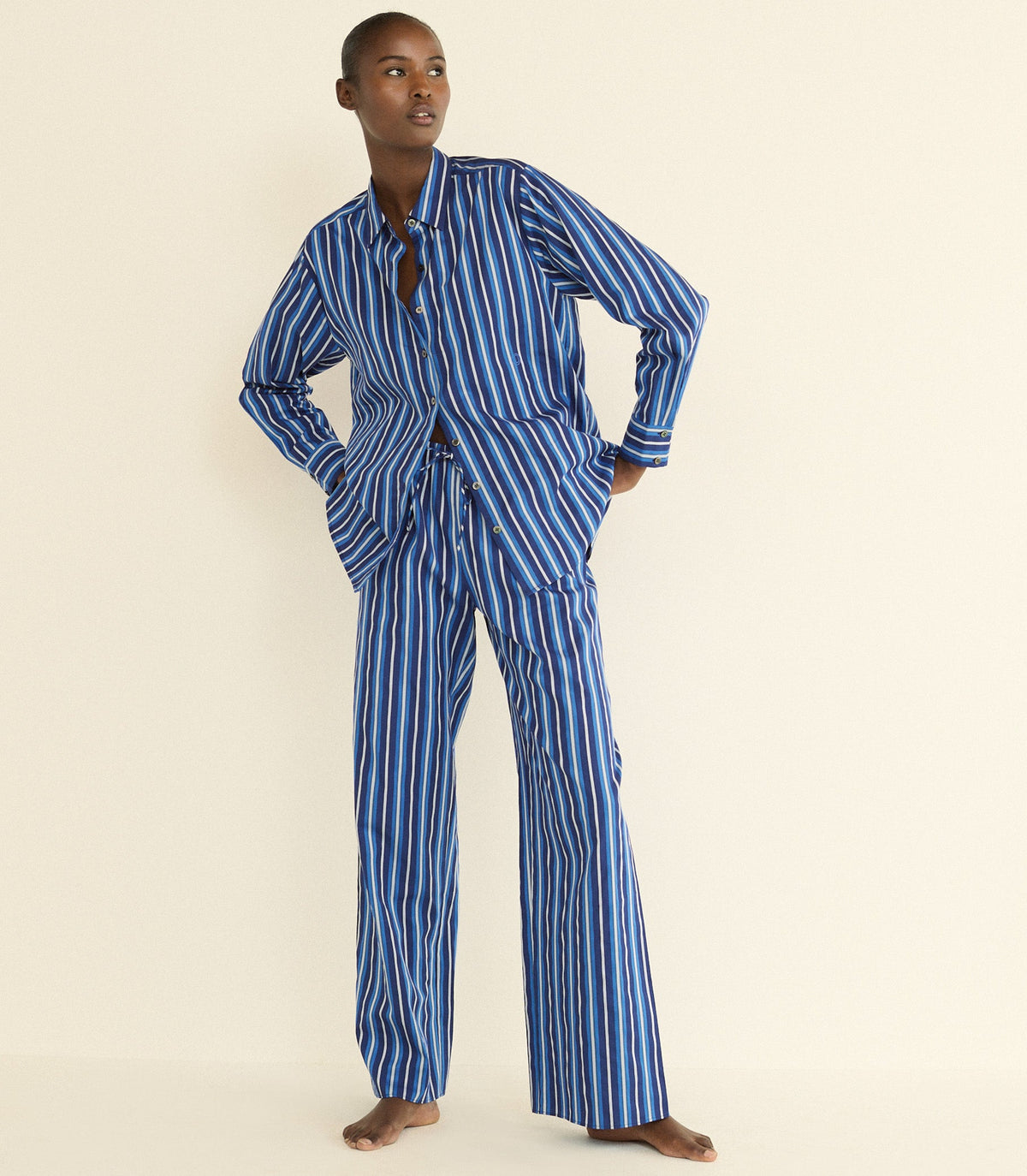 A model stands barefoot against a plain cream background wearing a matching two-piece pajama set with vertical blue and white stripes. Sporting a long-sleeve button-front top with a split neckline and wide-leg pants.