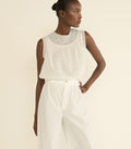 A model wearing a white sleeveless top with a detailed embroidered yoke and matching white wide-leg trousers, photographed against a solid light beige background.
