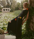 A woman in a black maxi dress with colorful flower appliqués leans against a vine-covered wall beside a lily pond.
