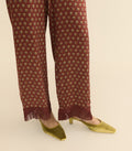 A close-up of a person's legs wearing rust-brown wide-leg trousers with a small floral pattern and fringe hems, paired with gold pointed-toe heels on a light beige background.