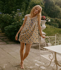 A woman stands barefoot on a stone patio in a garden setting, wearing a sleeveless floral babydoll dress with lace trim. A white metal garden table is visible to her right.